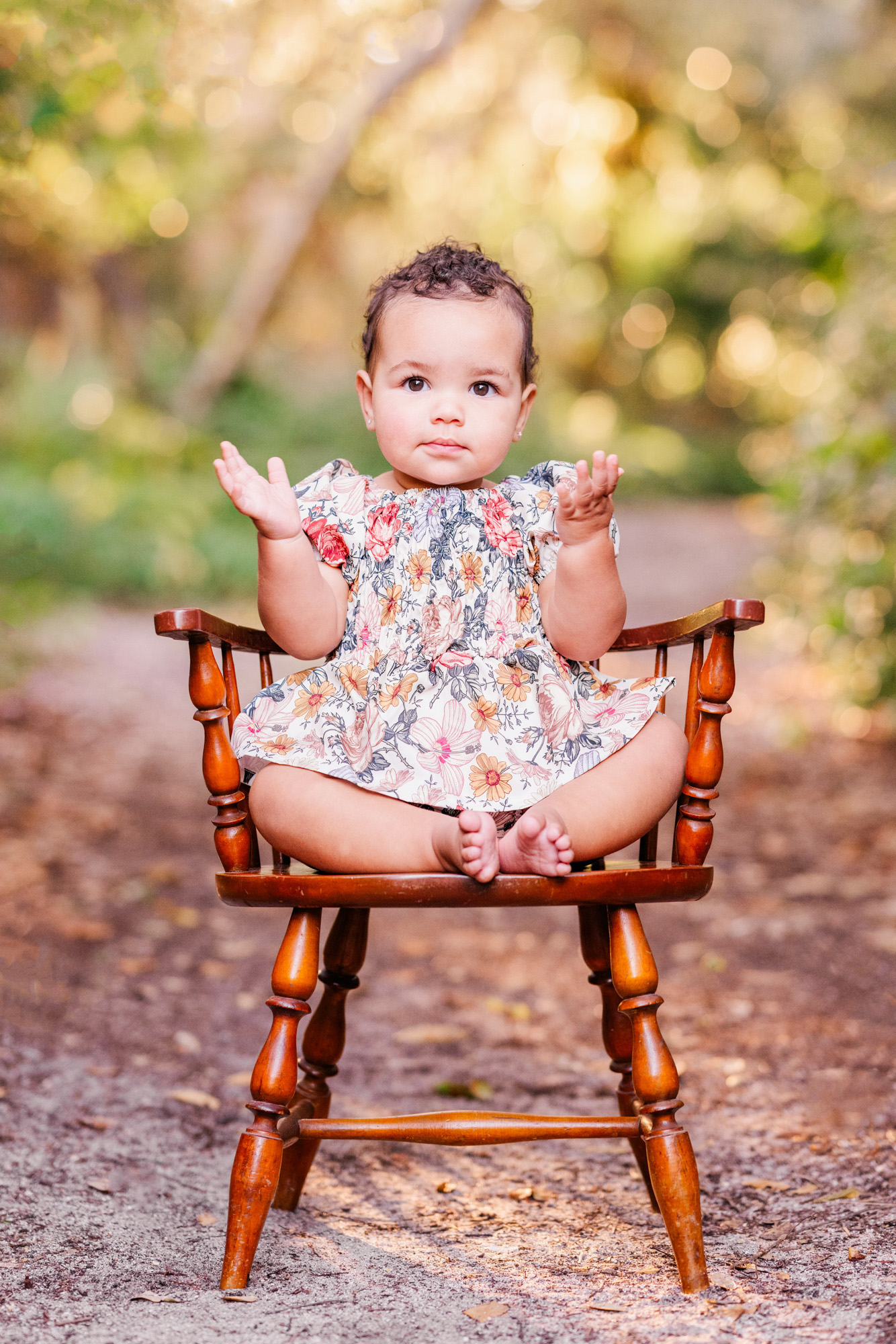 Baby raising both hands while sitting on a small wooden chair outdoors.