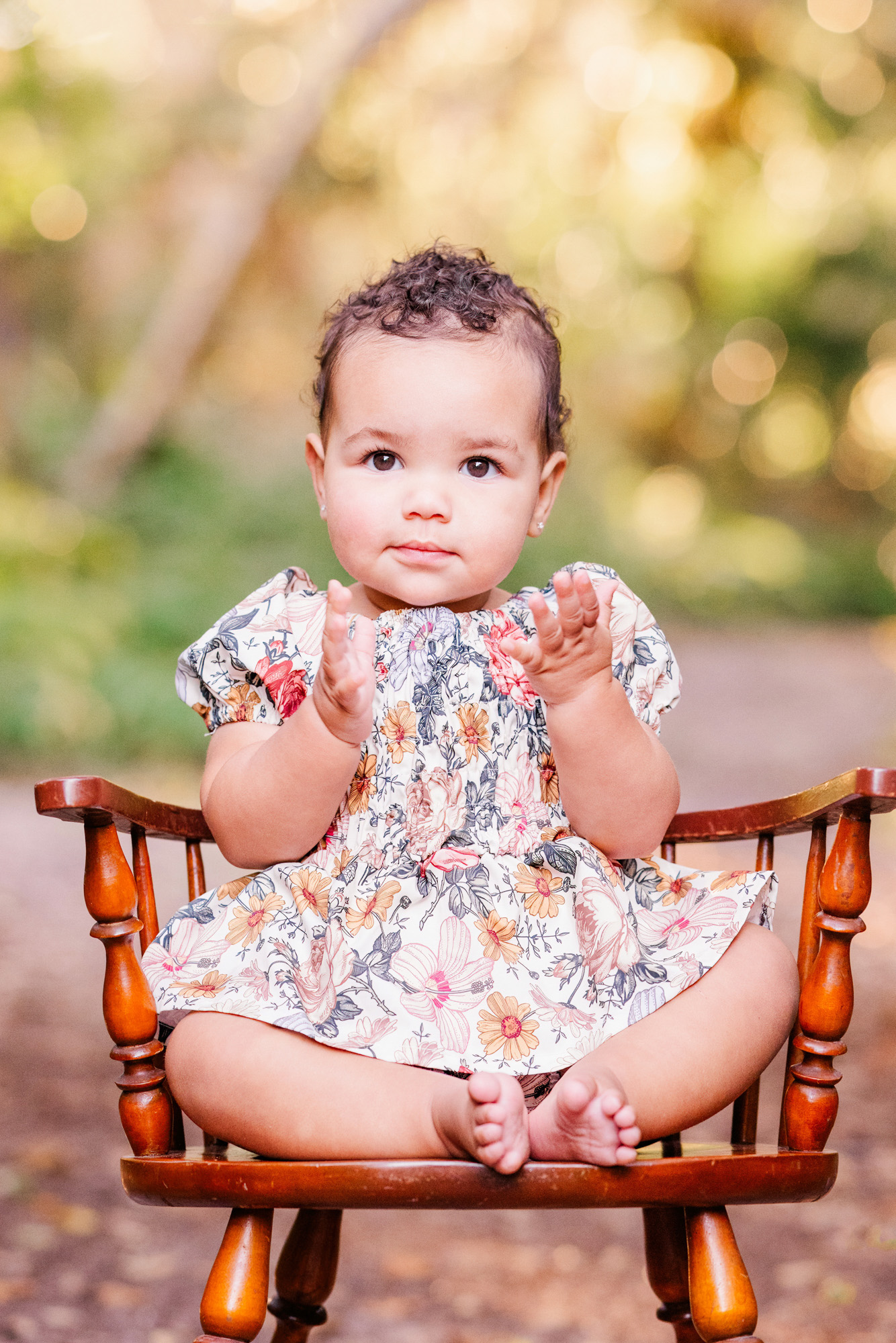 Close portrait of a baby sitting on a wooden chair with hands held together.