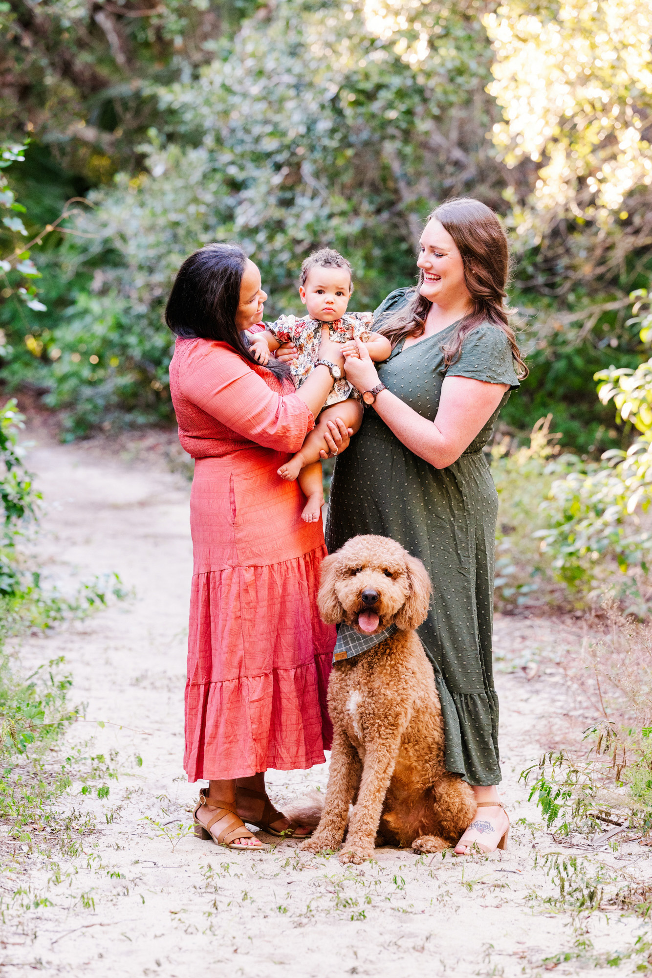 Baby looking toward the camera while both moms smile and hold them beside the family dog.