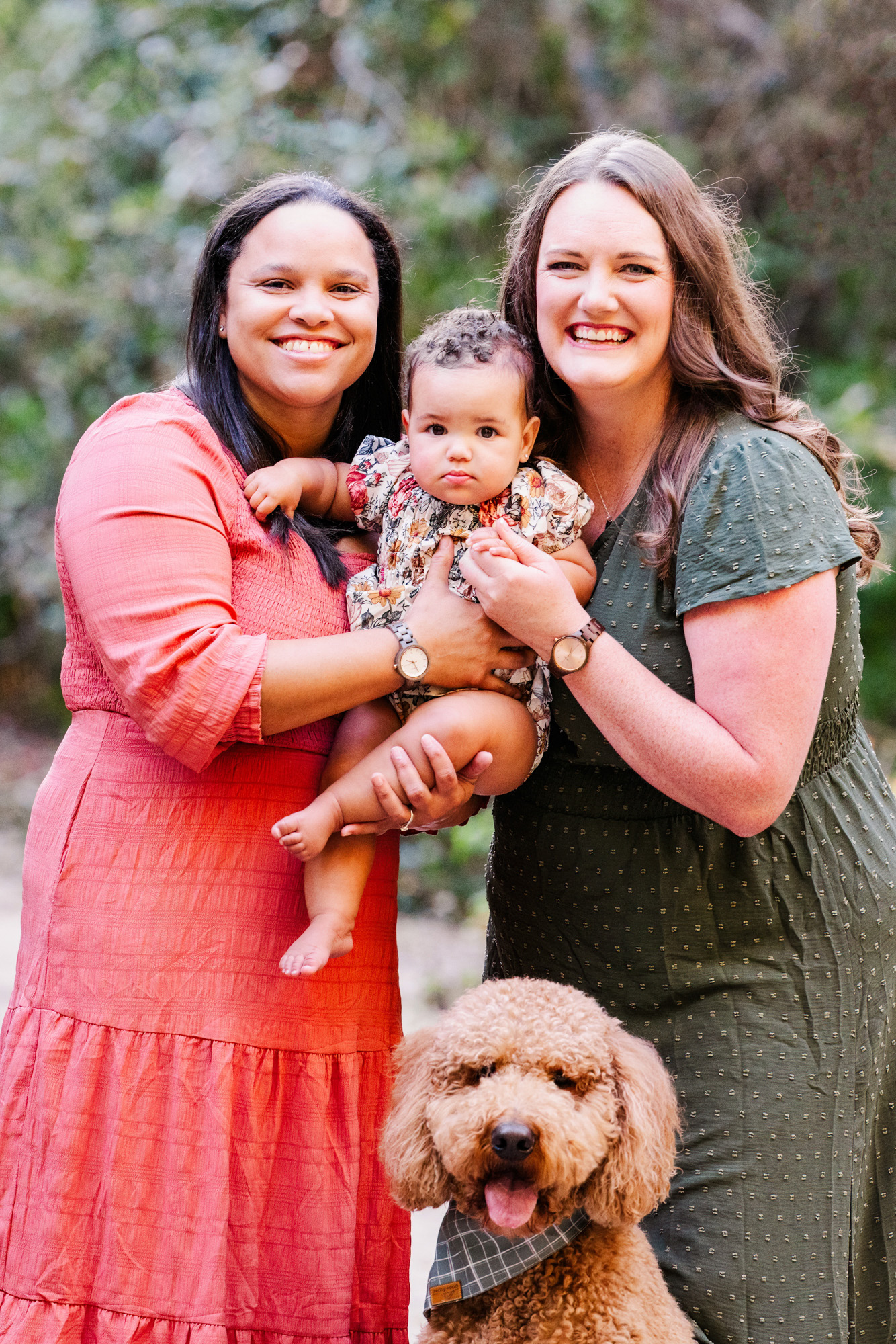 Two smiling mothers holding their baby together with the family dog in front.