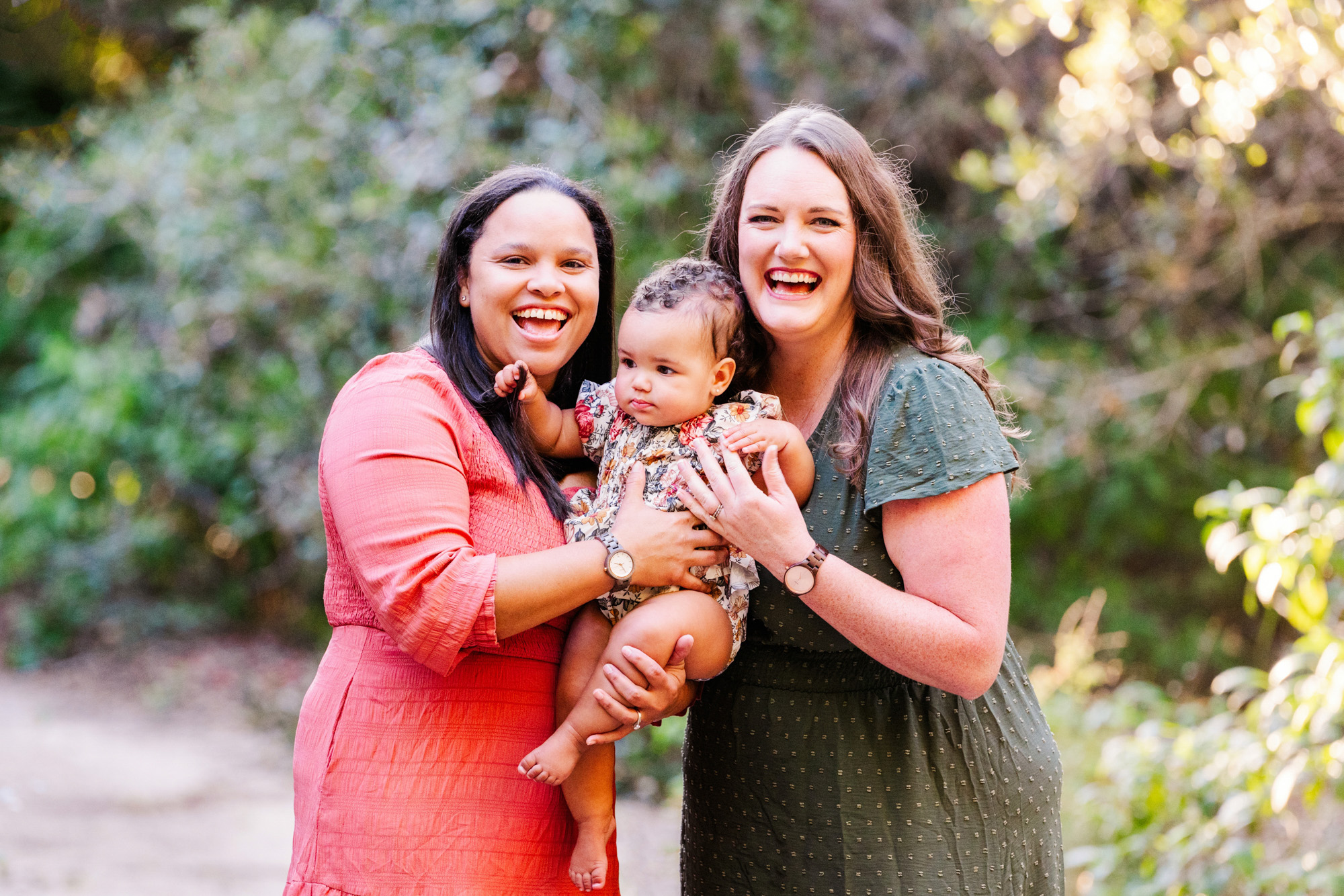 Two moms laughing while holding their baby during an outdoor family photography session.