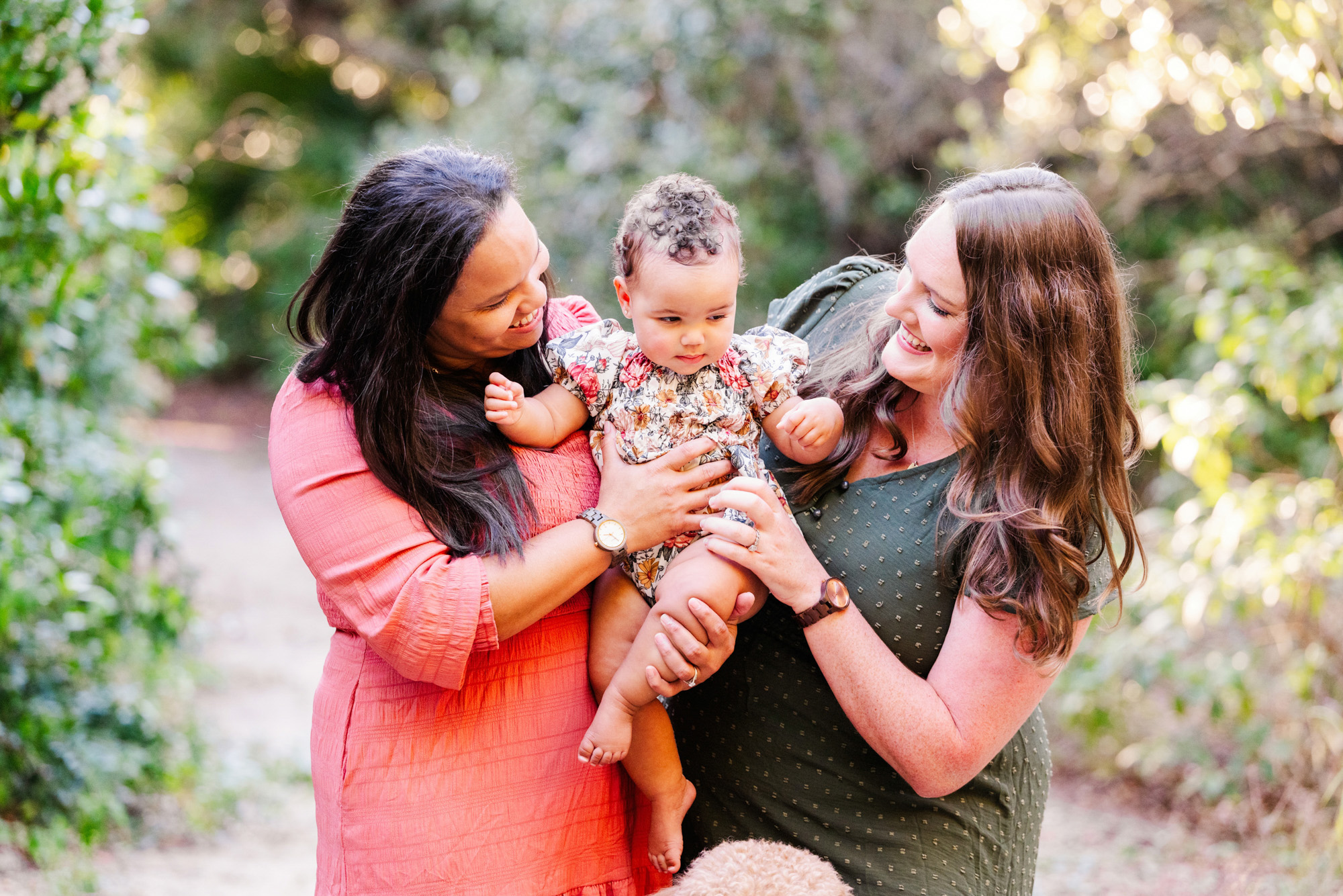 Two mothers looking lovingly at their baby surrounded by soft green foliage.