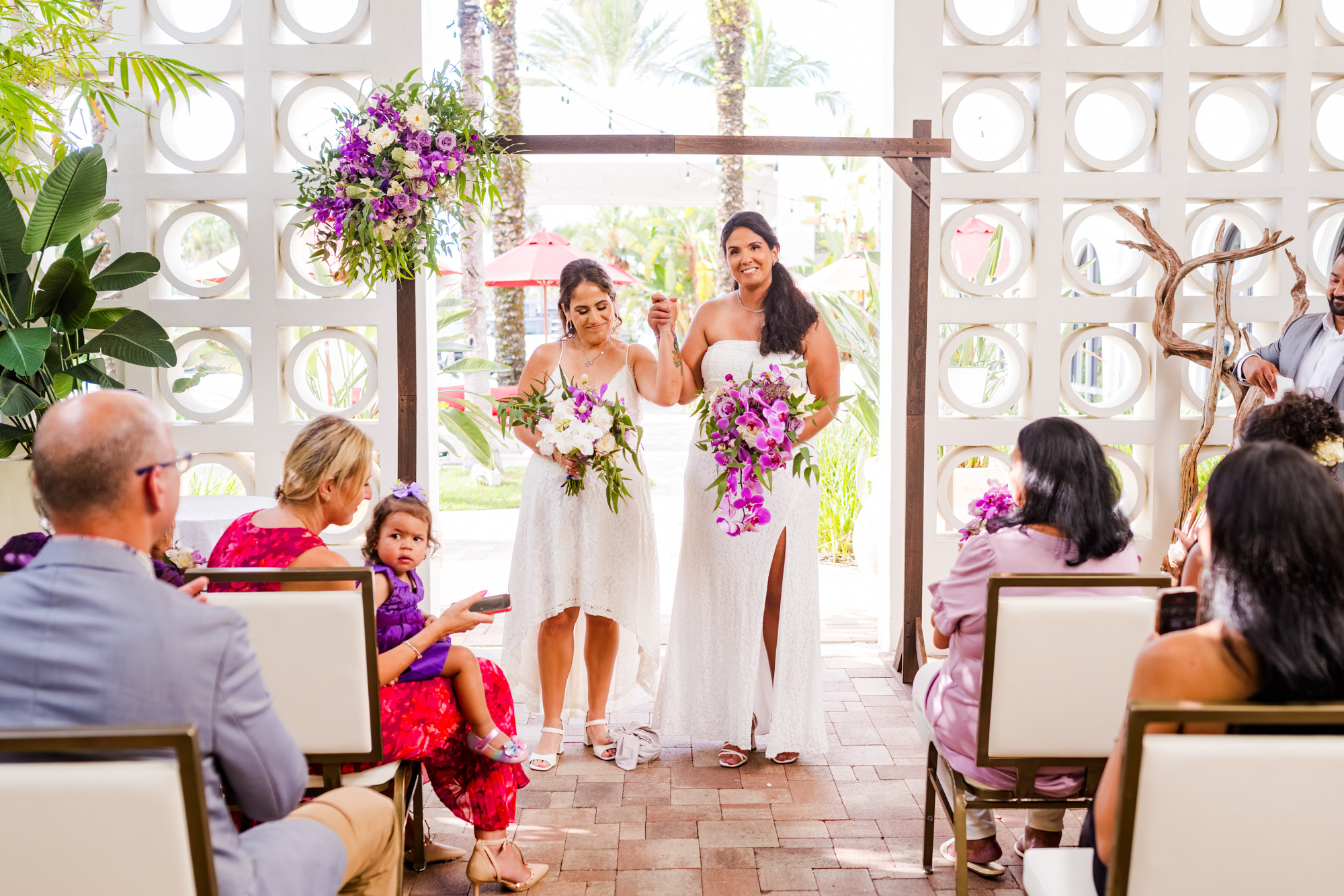 Two brides standing hand in hand beneath a simple wooden floral arbor during their wedding ceremony at Costa d'Este in Vero Beach