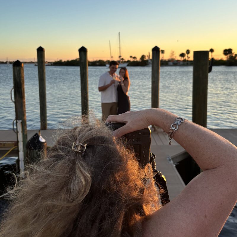 Behind-the-scenes view of Liz Cowie photographing a newly engaged couple at Eau Gallie Yacht Club at sunset.