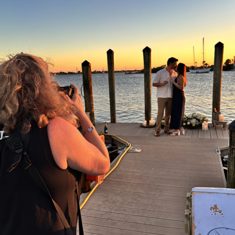 Behind-the-scenes view of Liz Cowie photographing a newly engaged couple at Eau Gallie Yacht Club at sunset.