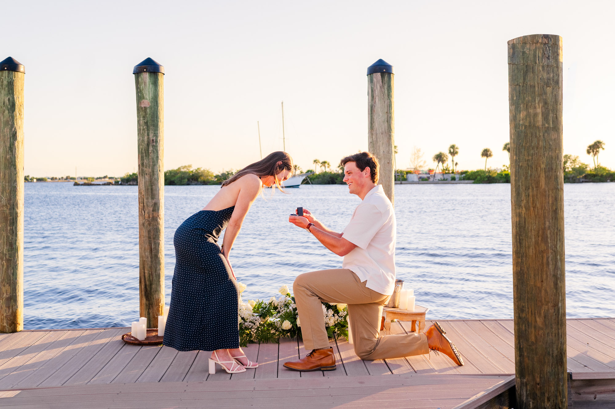 Woman leans forward with excitement as her boyfriend proposes on one knee beside the water.