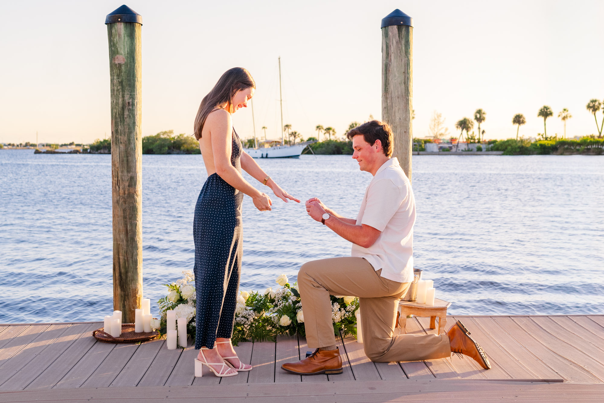 Woman smiles with her hands clasped as her boyfriend proposes against the waterfront sunset.