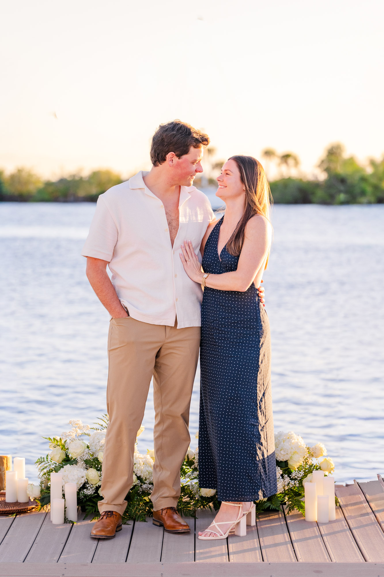 Couple sharing a quiet moment beside white floral arrangements and candles following their waterfront proposal.