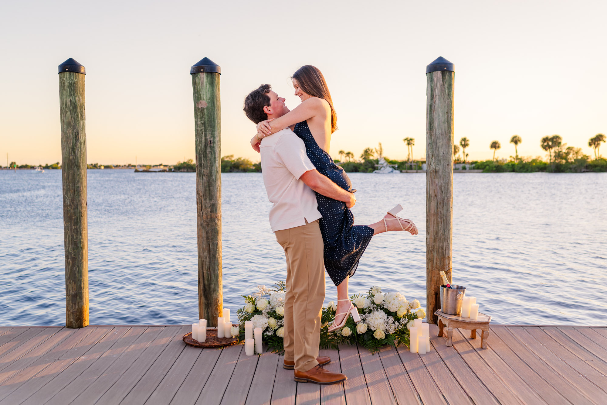 Man lifting his fiancée into his arms on a marina dock surrounded by white proposal flowers and candles.