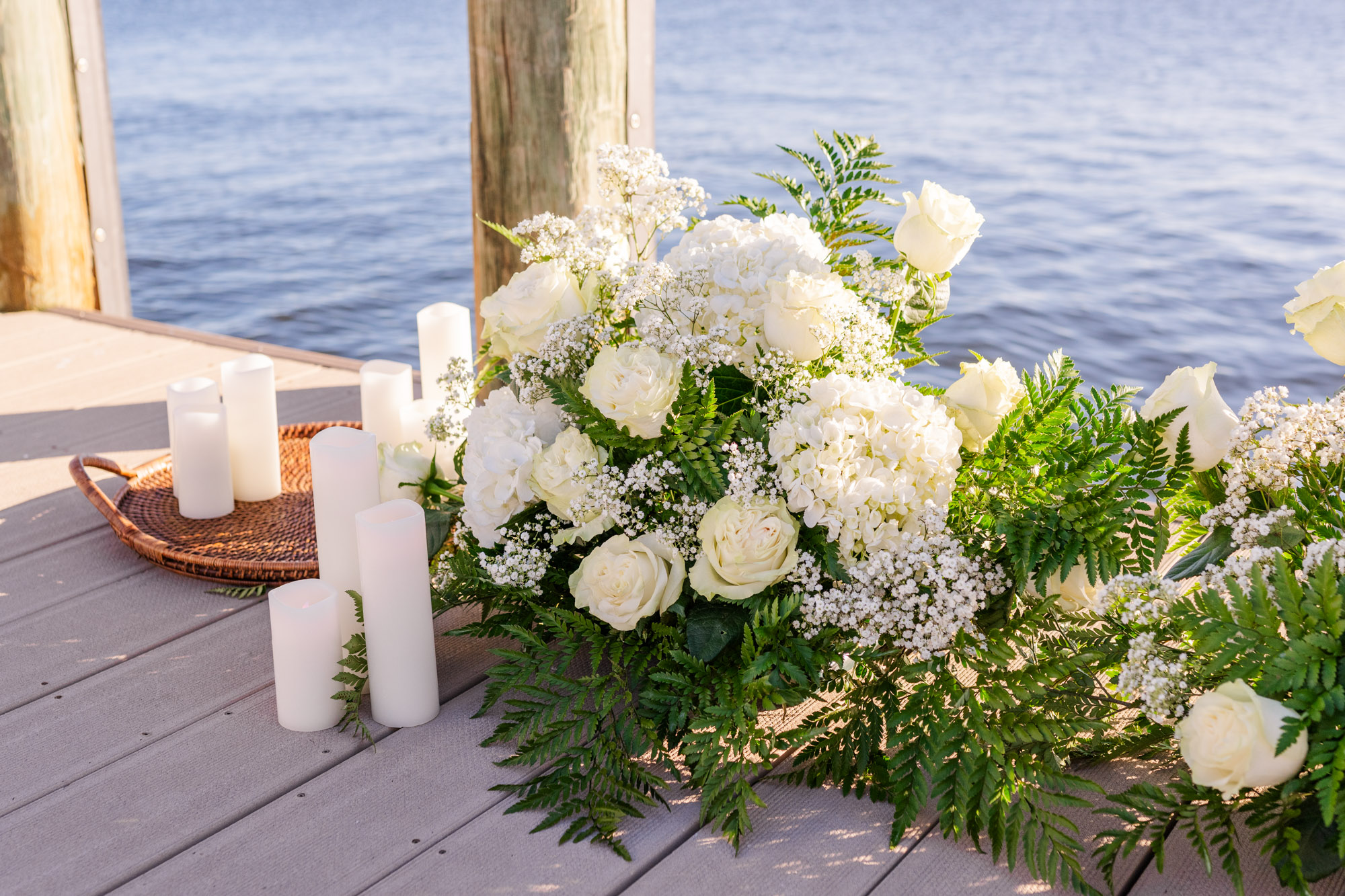 White roses, hydrangeas, baby's breath, and greenery arranged beside candles for a waterfront marriage proposal.