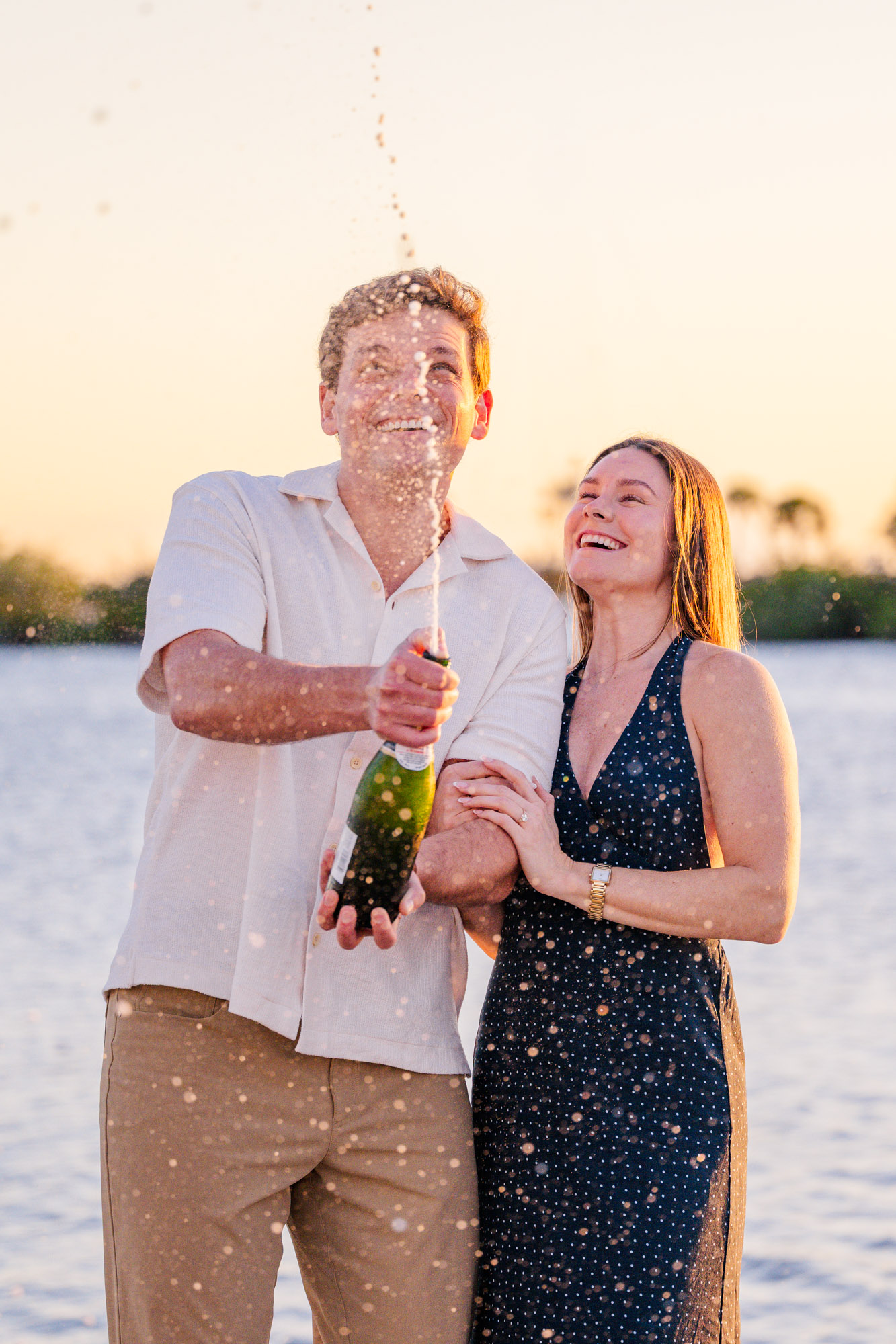 Man popping a bottle of champagne as his newly engaged partner laughs during their sunset proposal celebration.