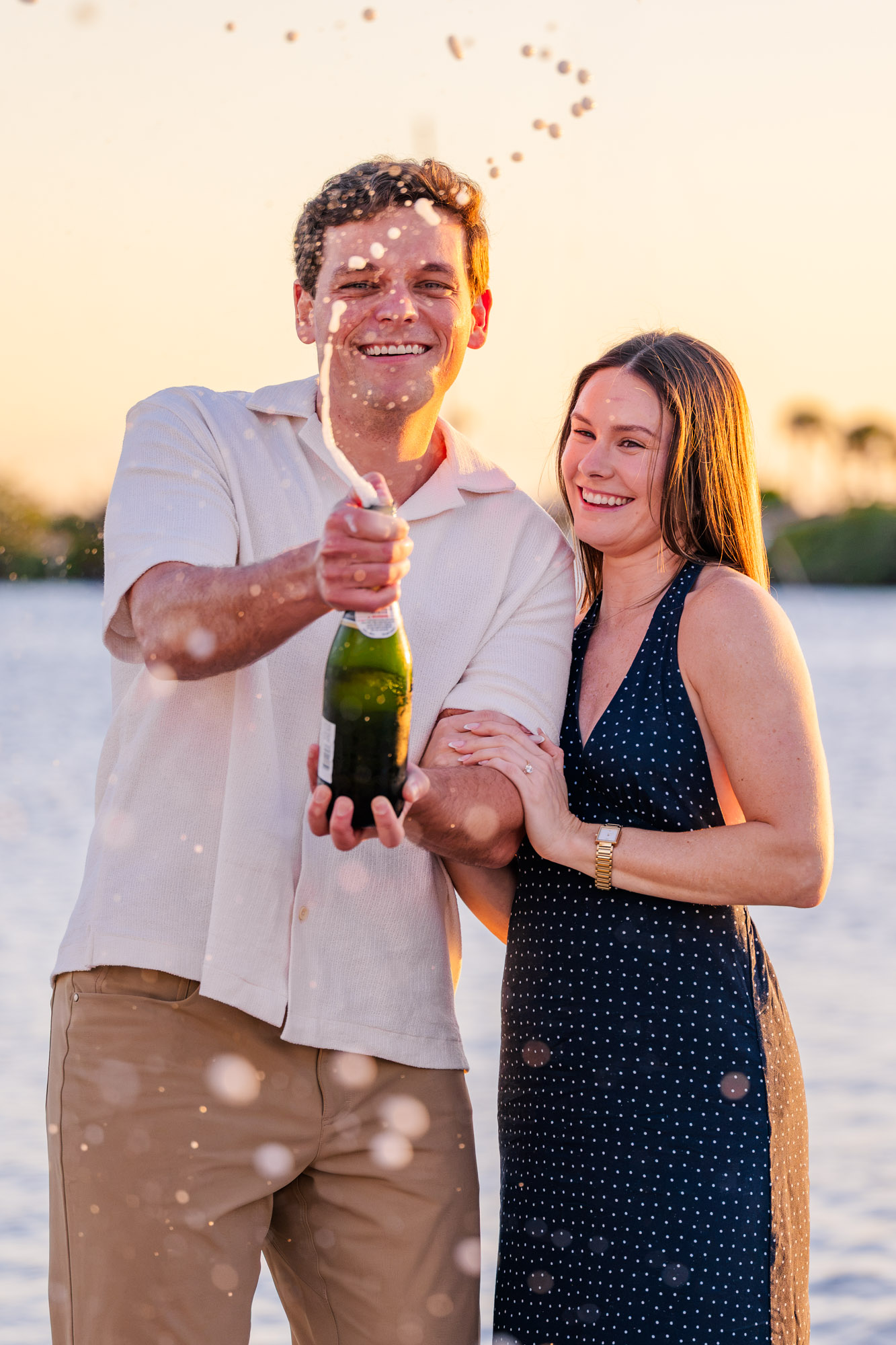 Champagne spraying through the air as a newly engaged couple celebrates their proposal beside the water.