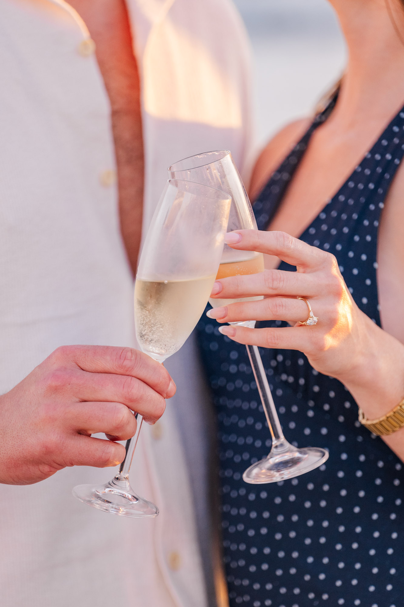 Close-up of two champagne flutes touching in a celebratory toast, with a diamond engagement ring visible.