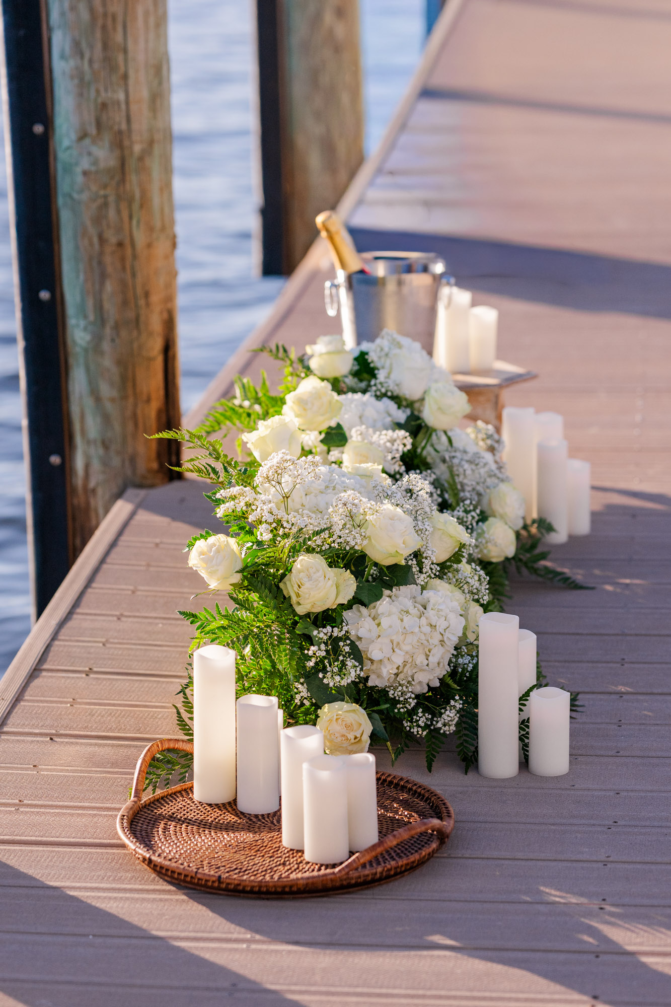 White floral arrangements, candles, and a champagne bucket arranged along a wooden dock for a sunset proposal.