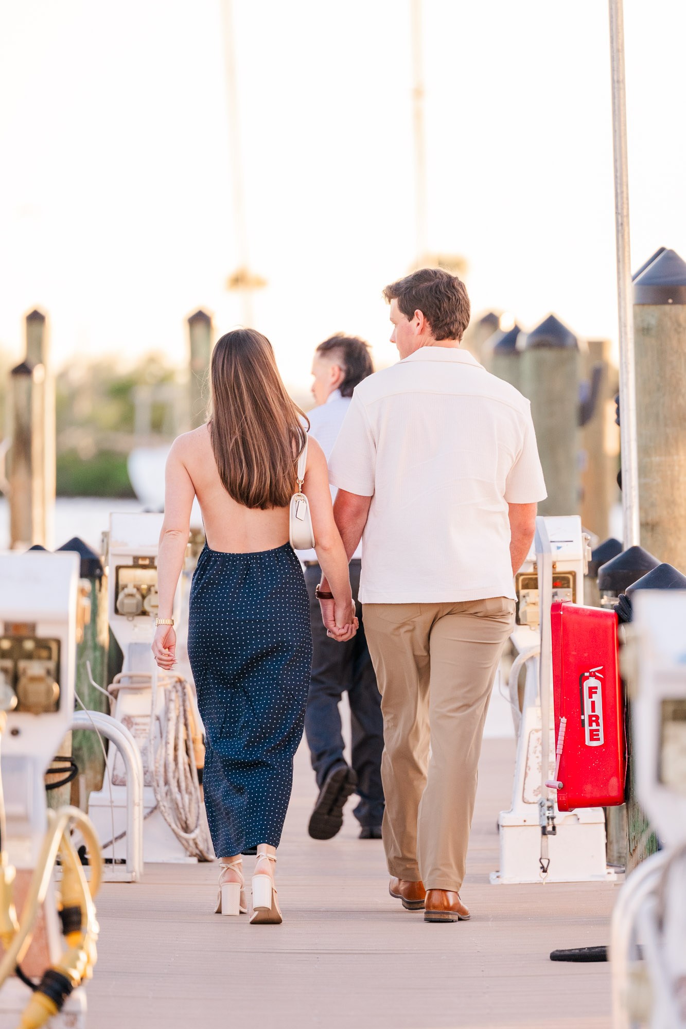 Couple holding hands as they walk along a dock lined with boats in the warm evening light.