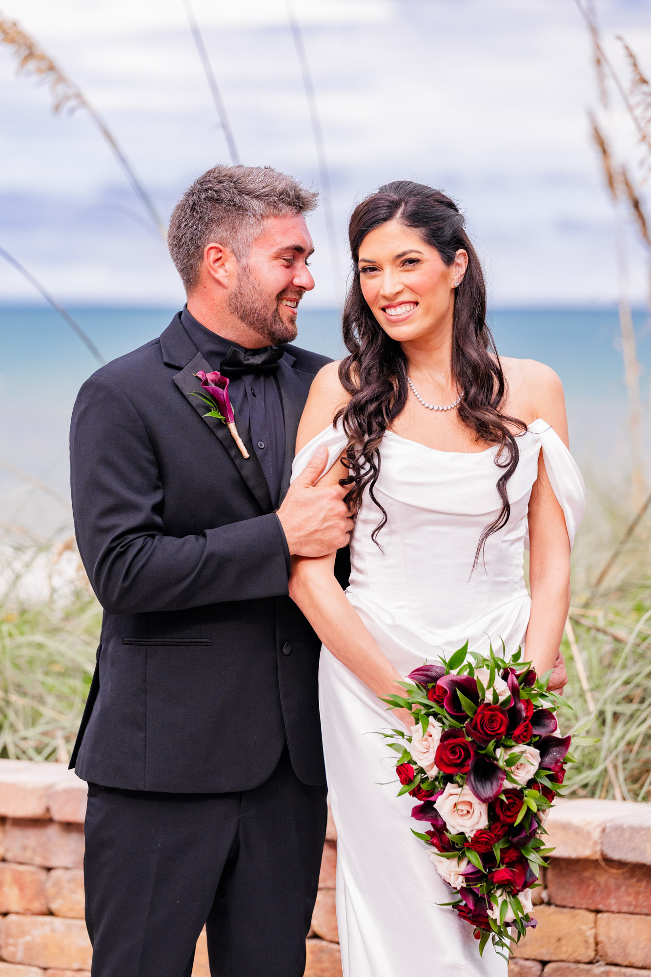 Groom laughing as he looks at his bride, who smiles at the camera holding a red and burgundy bouquet