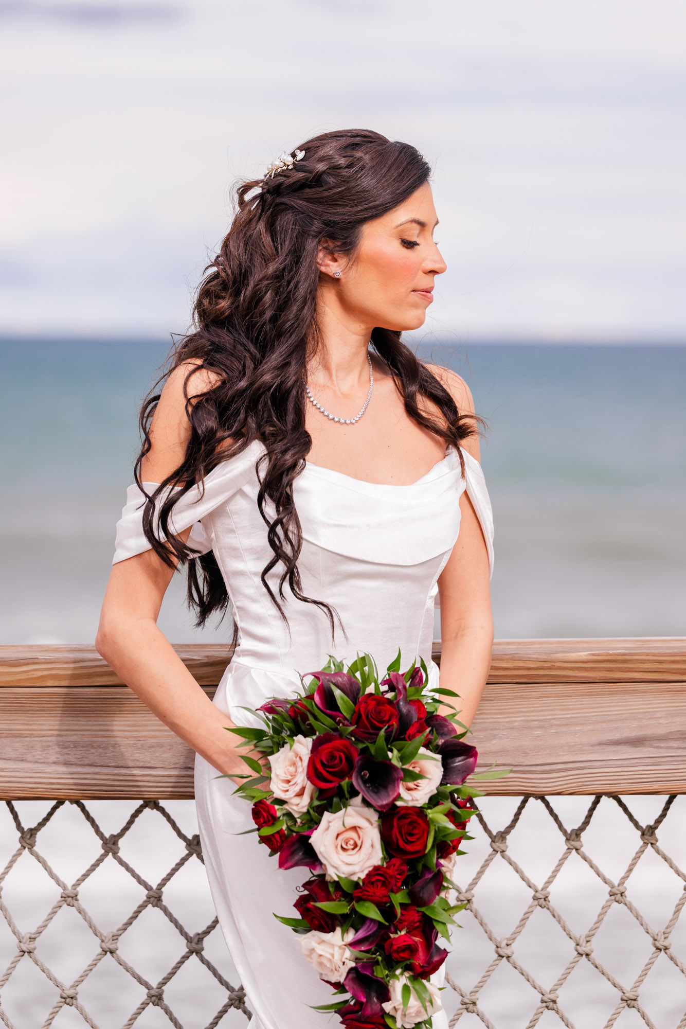 Bride in profile at the boardwalk railing with a cascading bouquet of red roses and calla lilies