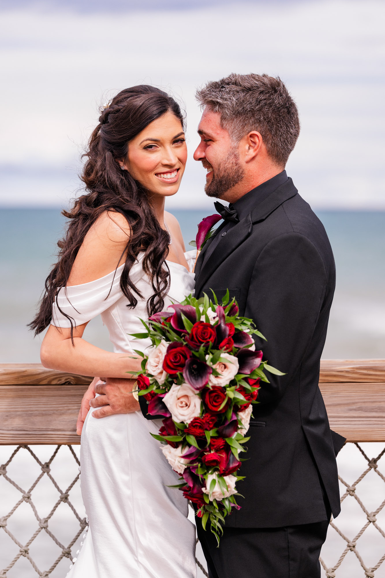 Bride and groom cheek to cheek on the boardwalk, both smiling, the Atlantic behind them