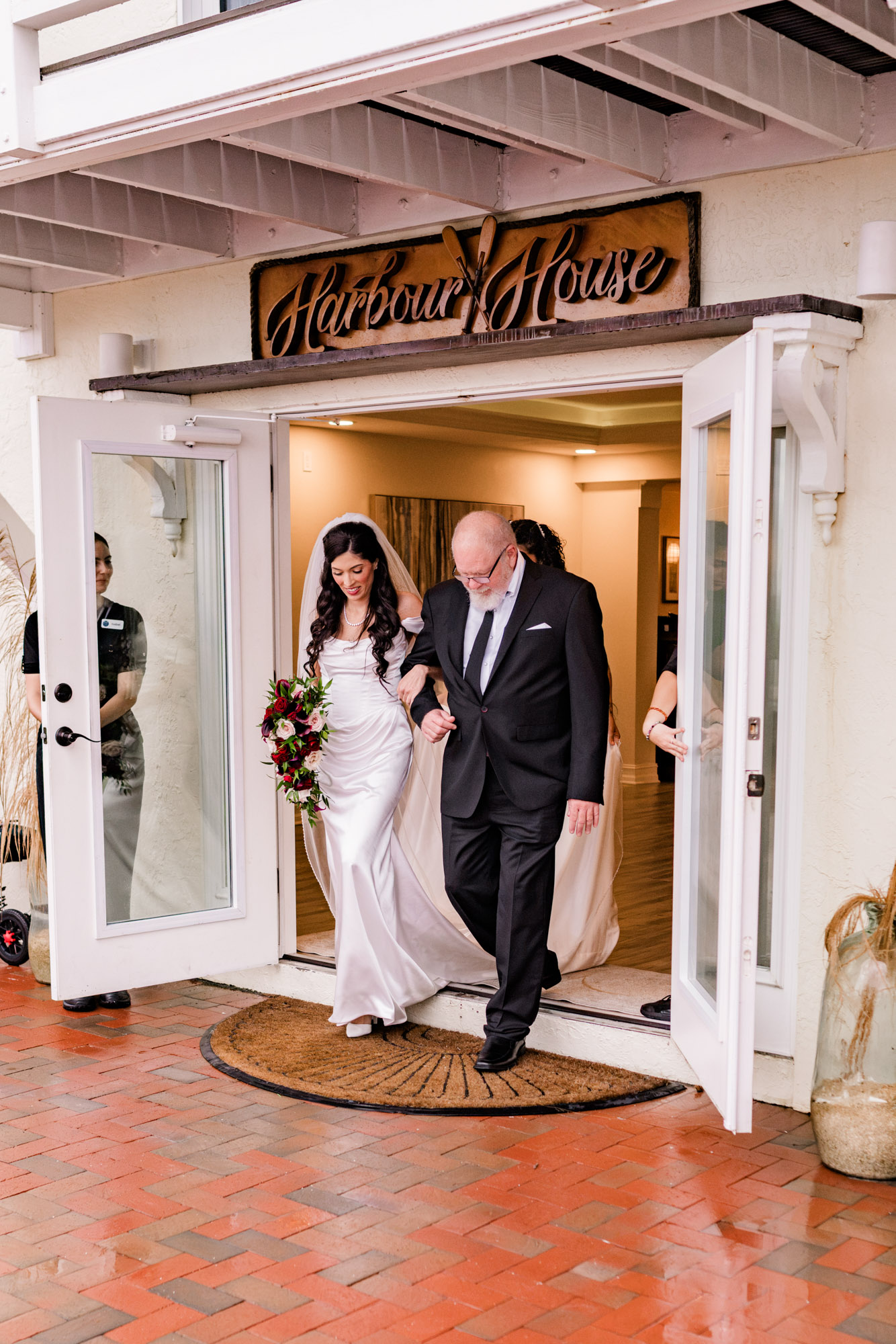 Bride steps out through the Harbour House doors on her father’s arm onto wet brick