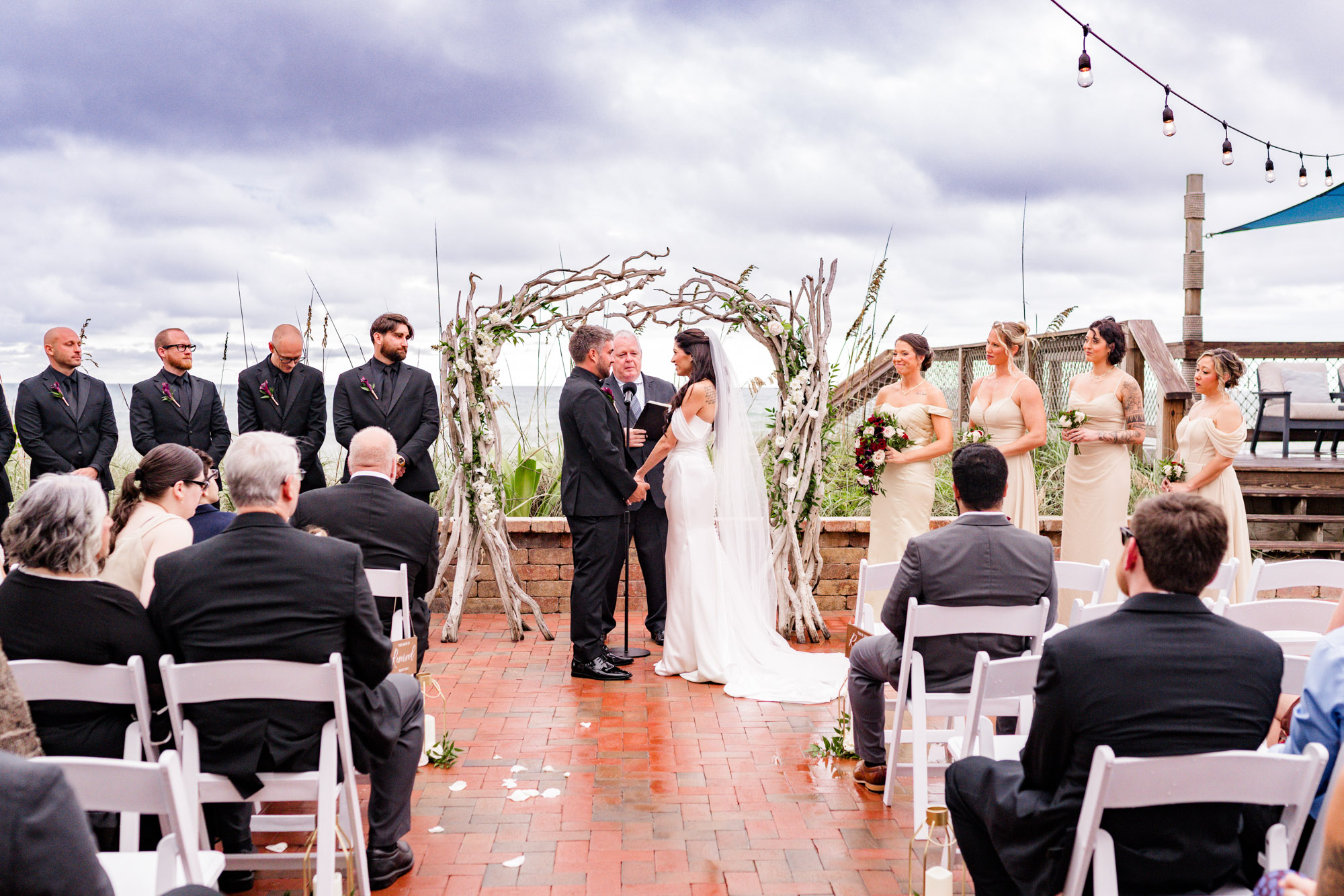 Bride and groom exchanging vows under a driftwood arch, wedding party either side and the ocean behind