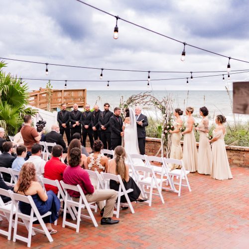 Wide view of the oceanfront ceremony, string lights overhead and guests seated on the brick patio