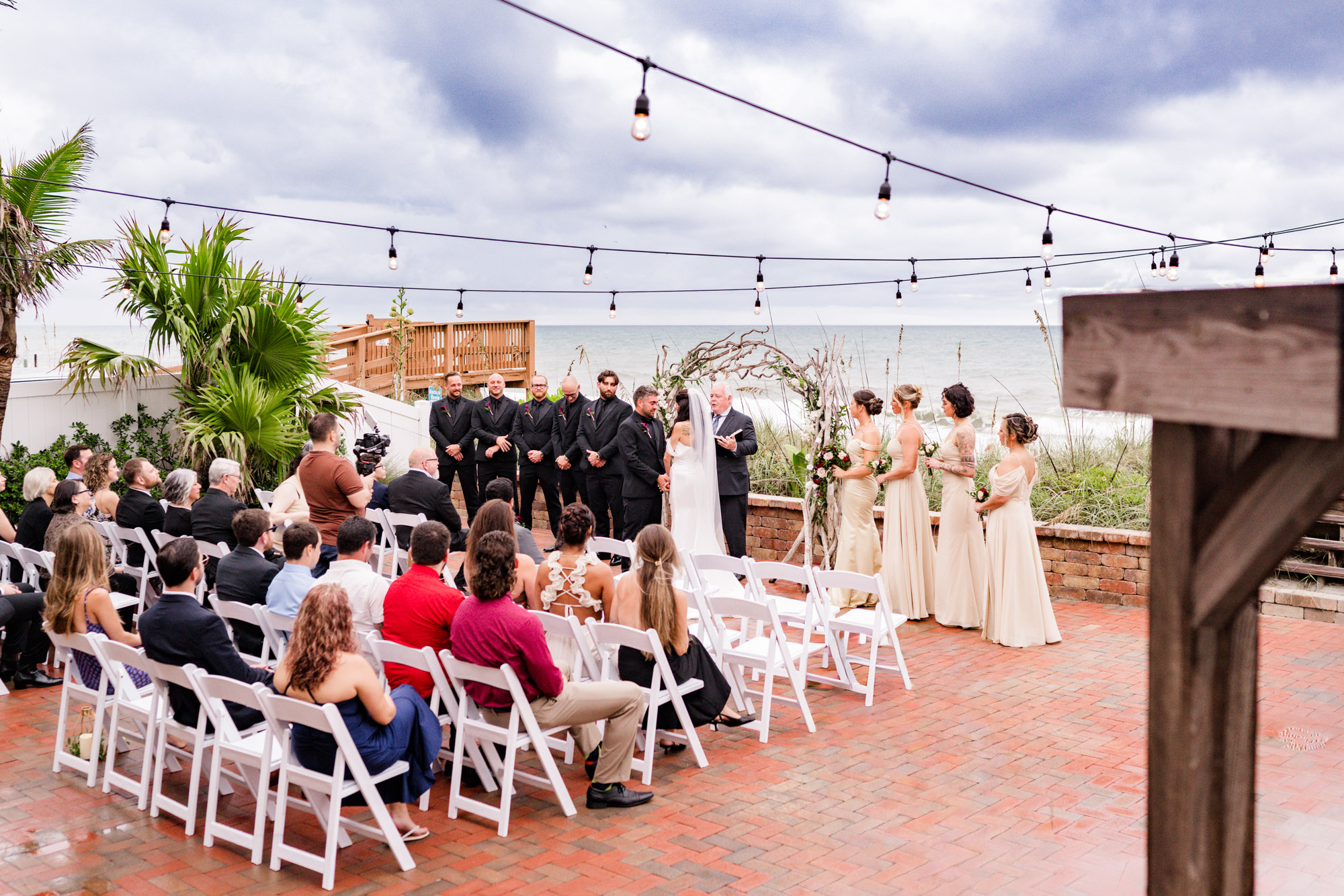 Wide view of the oceanfront ceremony, string lights overhead and guests seated on the brick patio