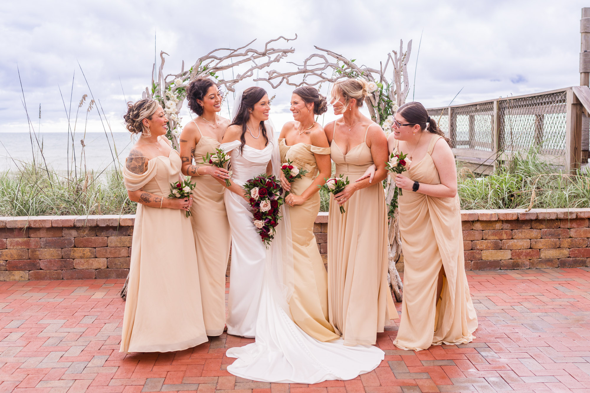 Bridesmaids turning to look at the bride, all laughing, the driftwood arch behind them