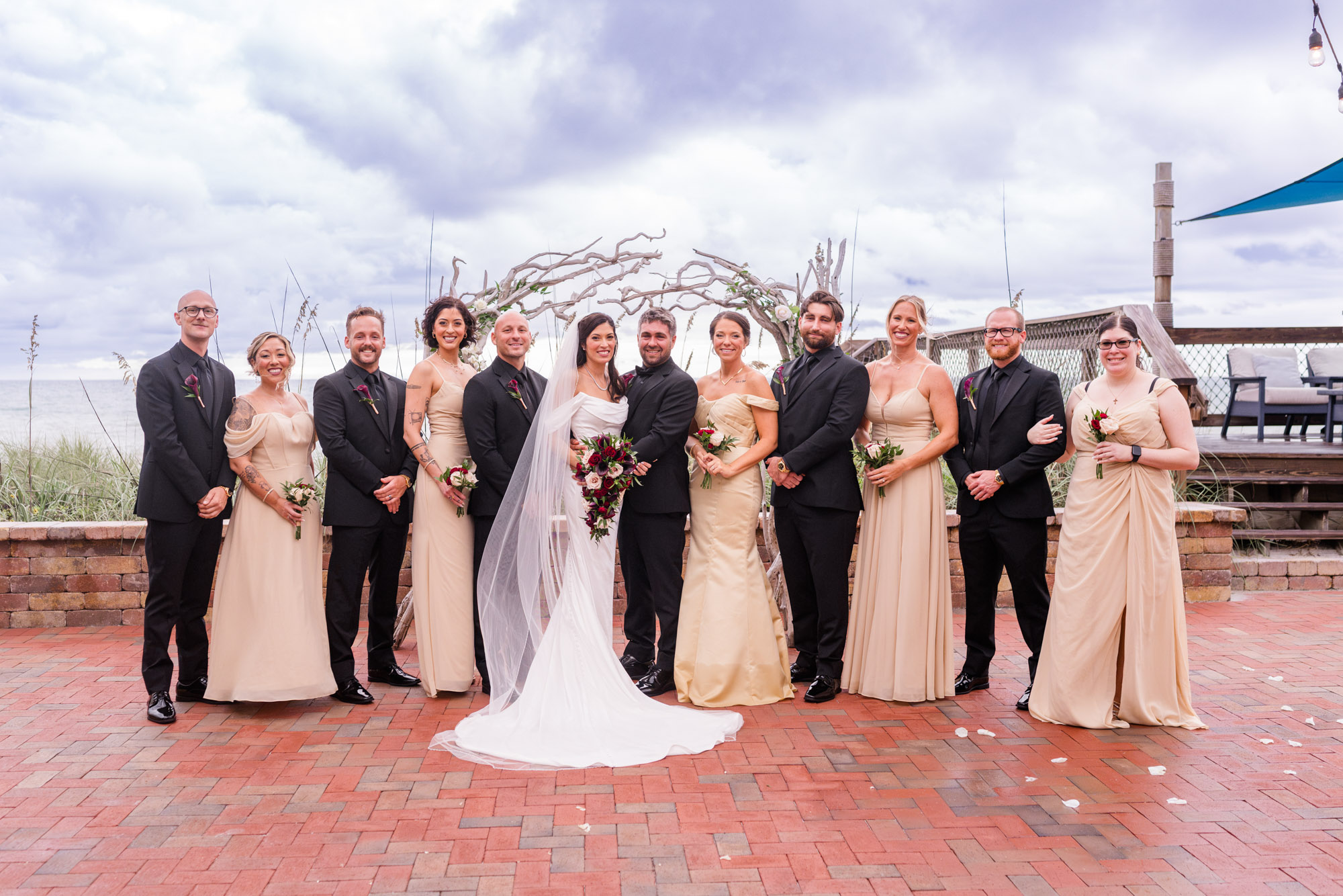 Full wedding party portrait under the driftwood arch, groomsmen in black and bridesmaids in champagne