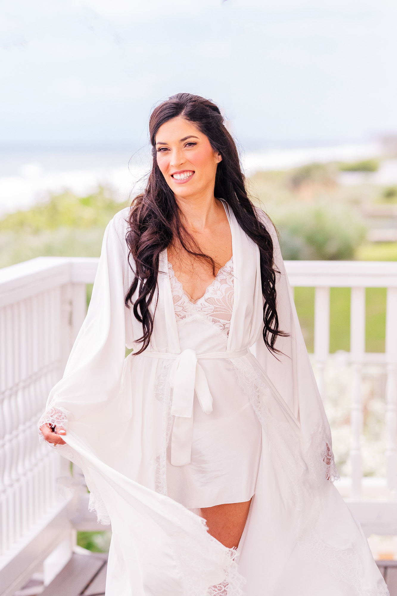 Bride leaning against a white deck railing in a lace robe, ocean behind her