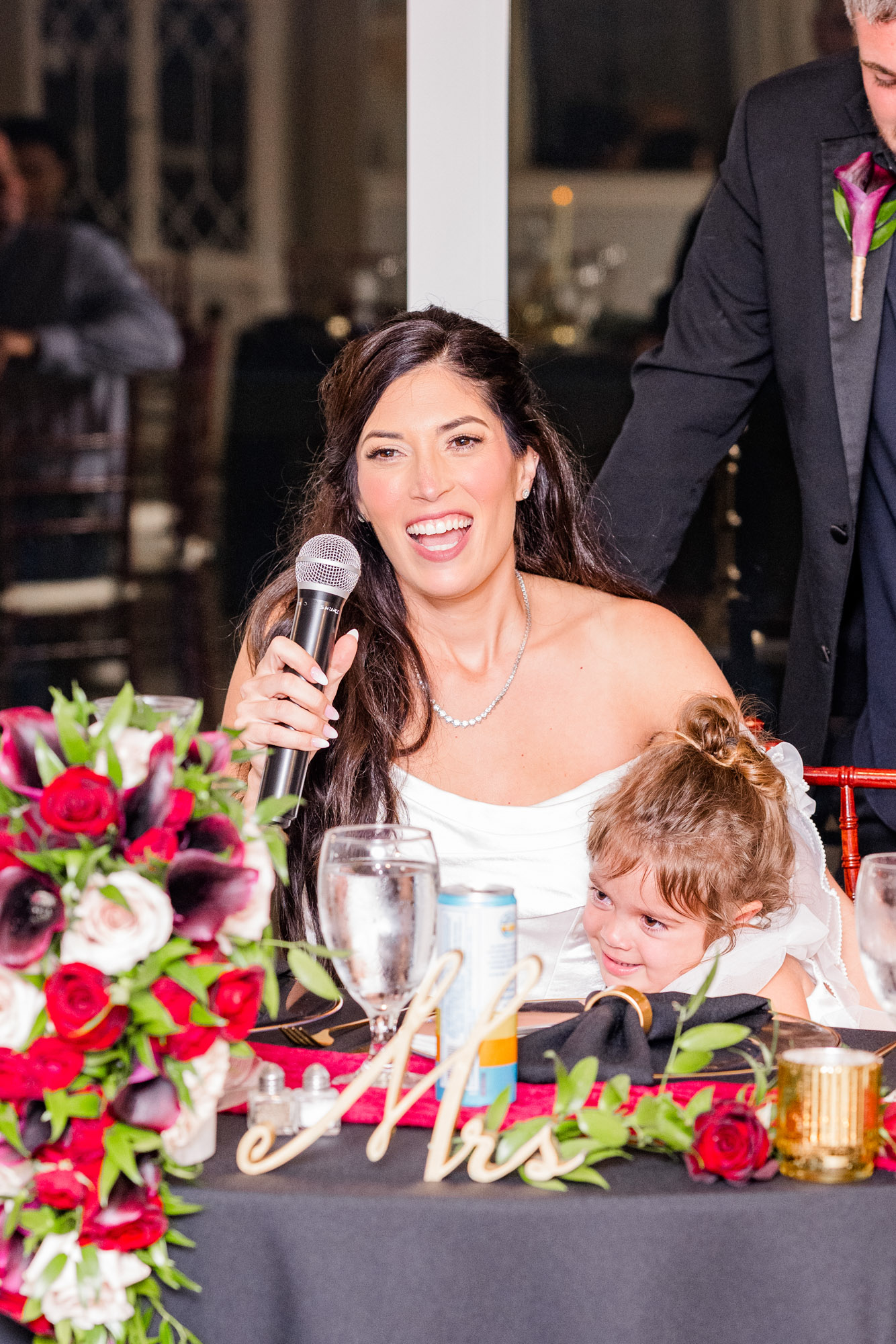 Bride laughing into a microphone at the sweetheart table while the flower girl leans against her