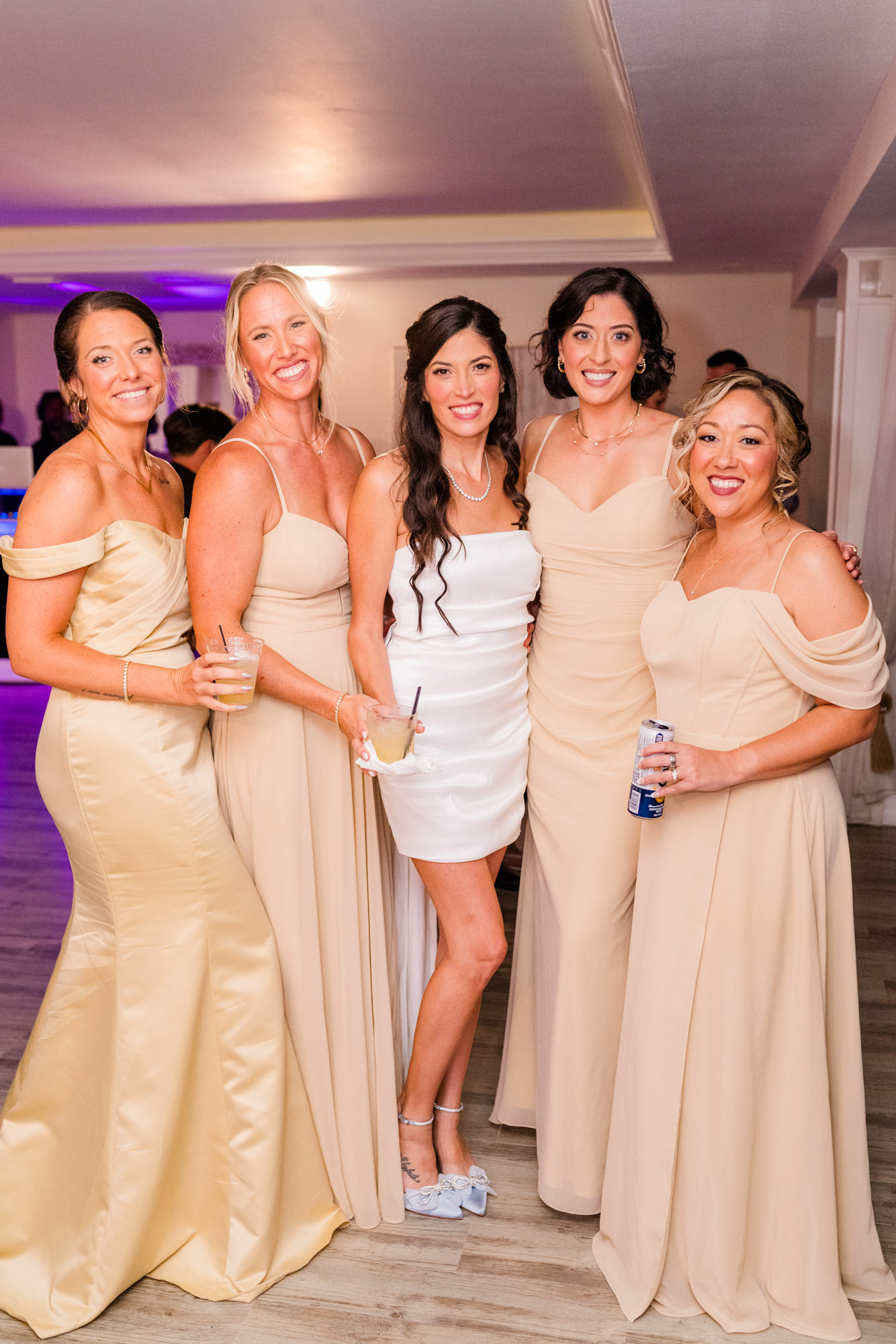 Bride and four of her bridesmaids at the reception, drinks in hand under purple uplighting