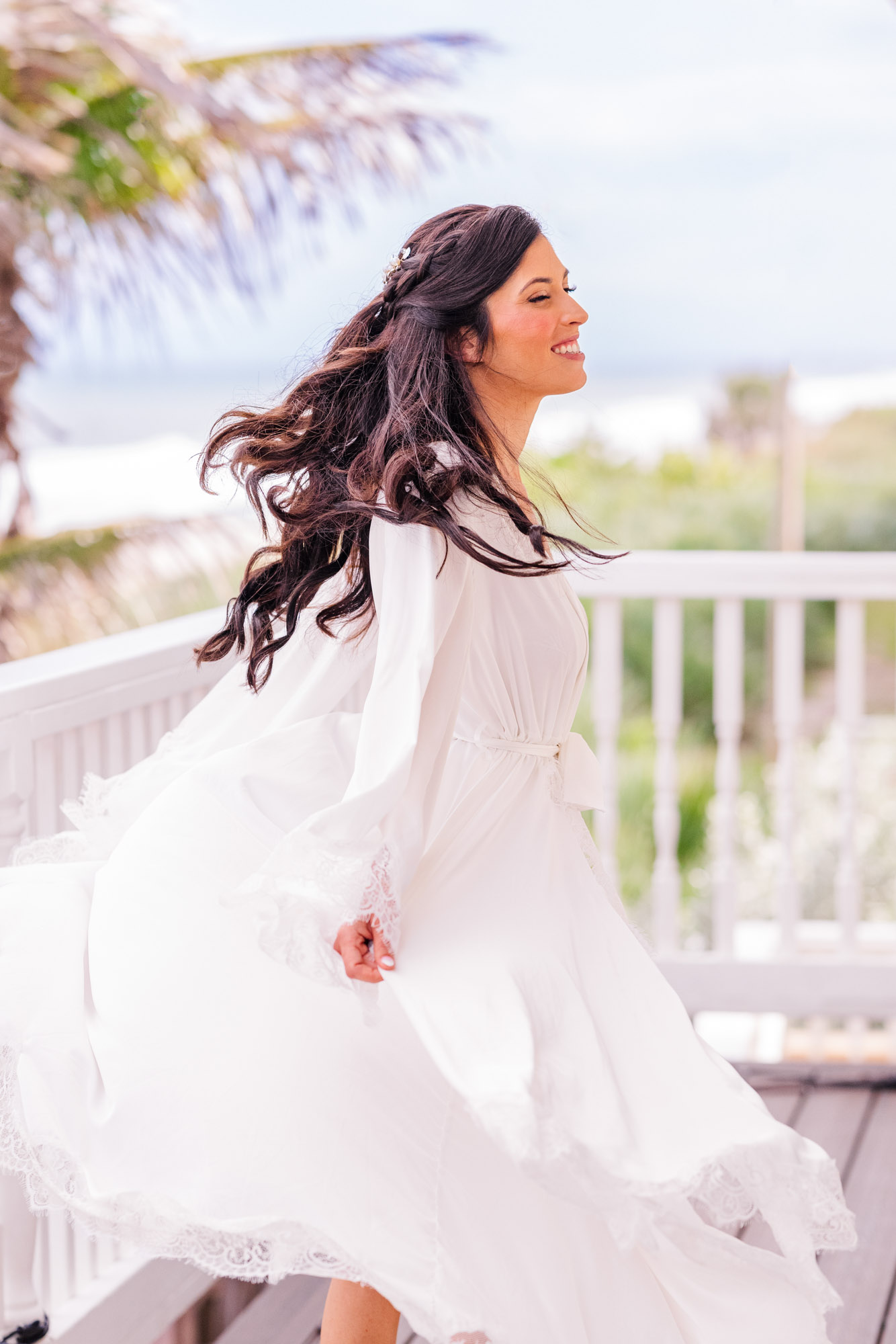 Bride laughing as she holds open a white lace robe on a deck overlooking the Atlantic