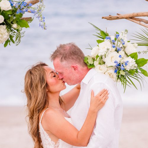 Close-up of newlyweds sharing a kiss beneath their blue and white floral wedding arch on Melbourne Beach.