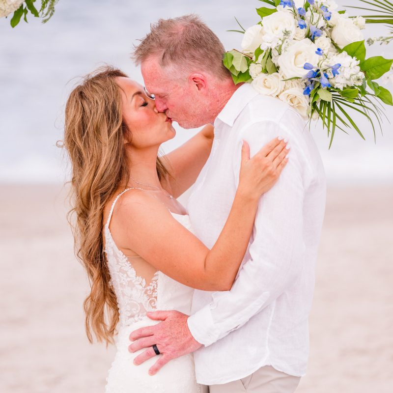 Close-up of newlyweds sharing a kiss beneath their blue and white floral wedding arch on Melbourne Beach.