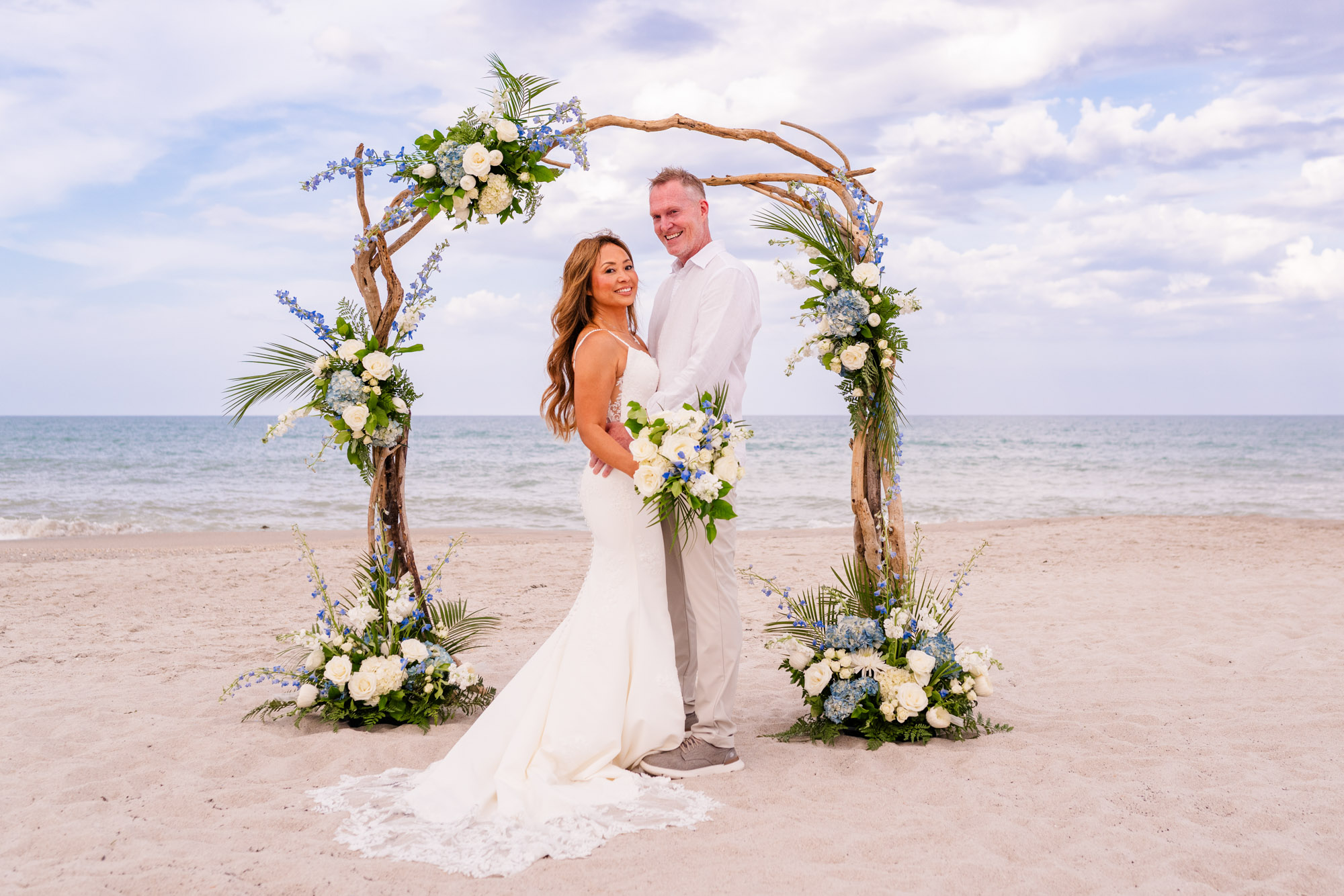 Bride and groom posing together beneath a rustic driftwood wedding arch decorated with blue and white flowers.