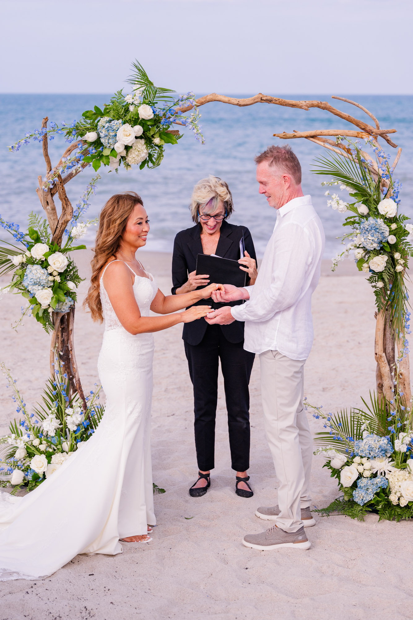 Couple exchanging wedding rings beneath a driftwood floral arch during their intimate beach wedding in Melbourne Beach, Florida.