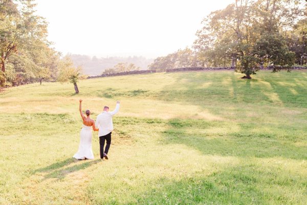 Bride and groom walking together through a glowing summer field in Pawling NYinto the sunset with their arms in the air